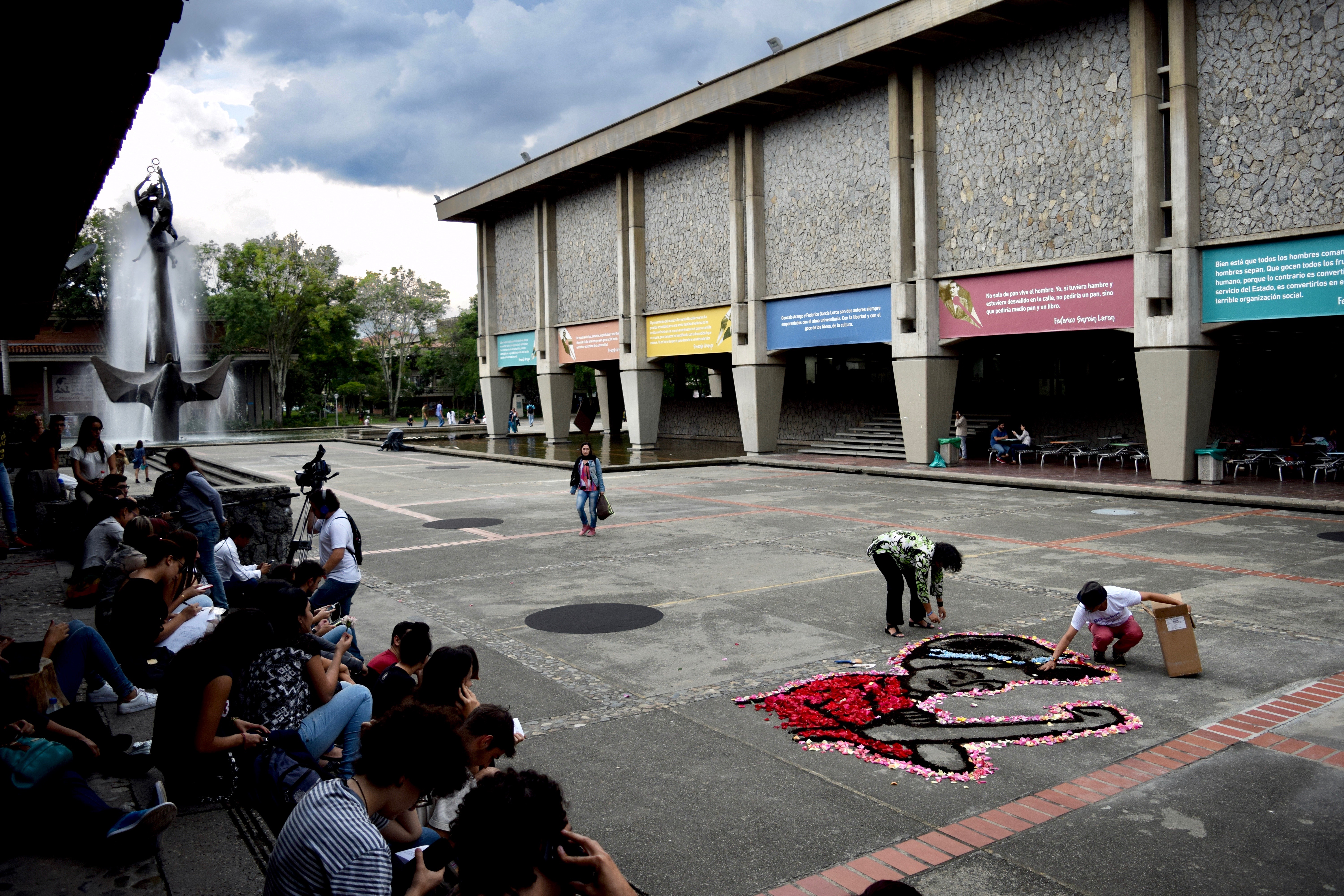 Personas reunidas frente a la biblioteca central UdeA, y dos personas realizando un performance de flores en suelo