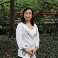 Fotografía de la profesora Ángela Milena. Plano medio. Tiene una camisa con bordados y al fondo árboles.
