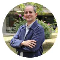 Profesor Esteban López Zapata, de traje azul, sonriente y con los brazos cruzados, posando al aire libre frente al Bloque 13 de la UdeA