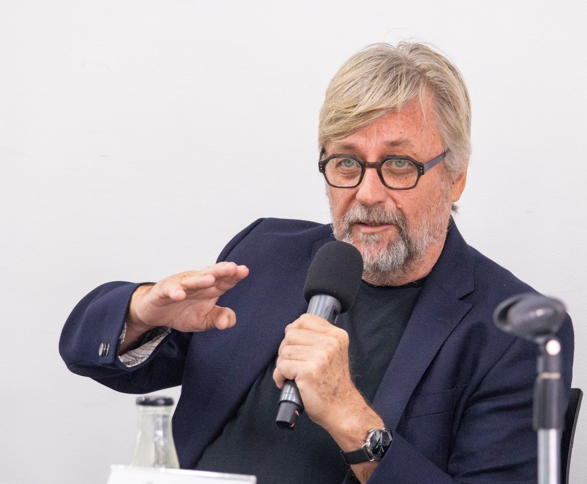 Scott O’Neill en conferencia, hablando con un micrófono. Está vestido con un traje azul marino y una camisa negra. Su mano derecha está extendida hacia el público, como si estuviera haciendo un gesto de invitación o aclarando un punto.